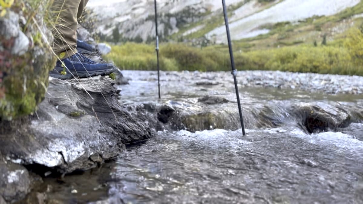 Hiker crossing a shallow stream with trekking poles and hiking boots.